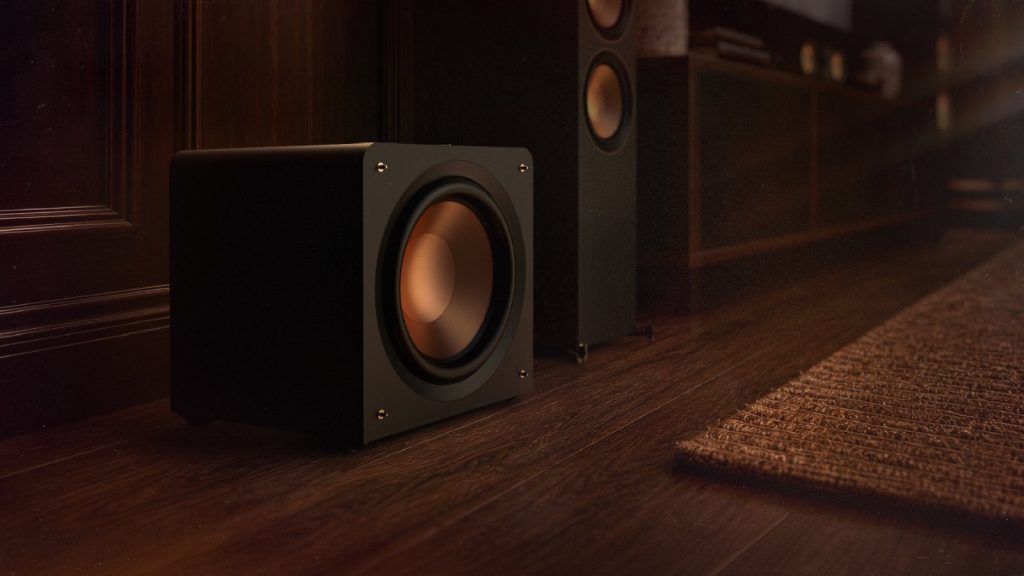 Klipsch Reference Premiere compact sealed subwoofer on the floor beside a media console and floor-standing speakers